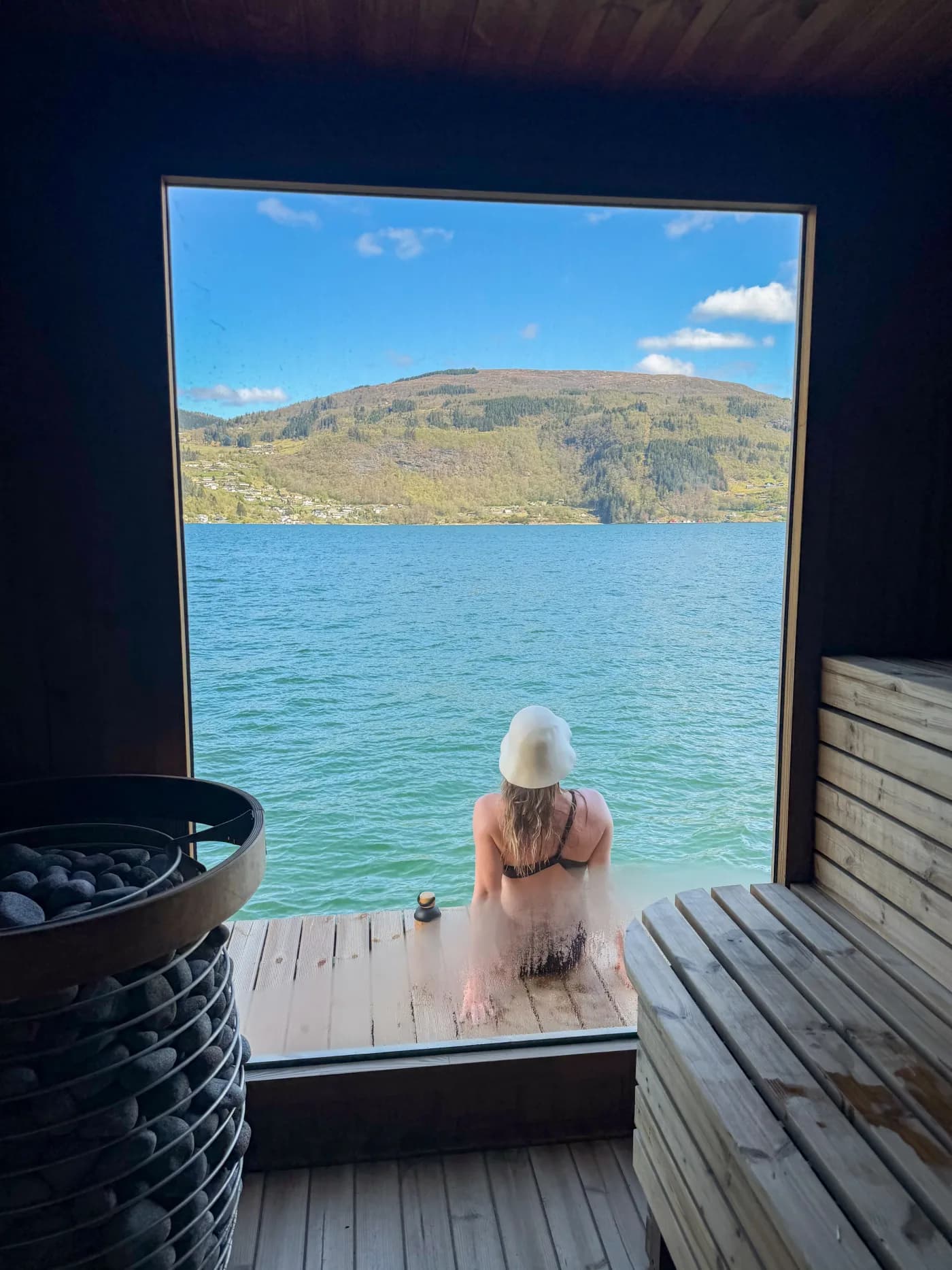 En badegjest i badstuhatt sitter i døråpningen til Ytre Arna Sauna og ser ut over fjorden
