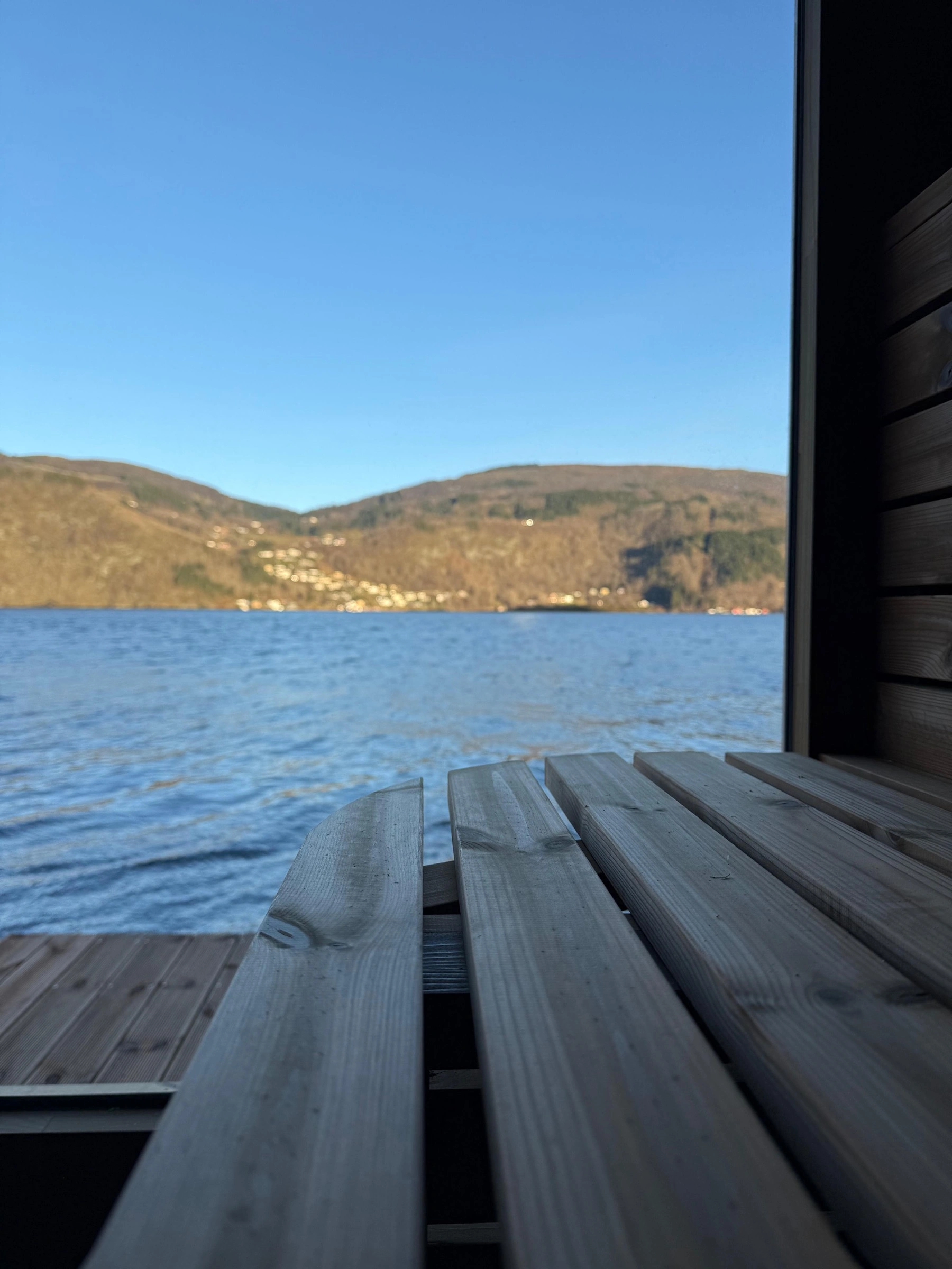 Utsikt over Vestfjorden gjennom åpningen i en badstu på bryggen — blå himmel, fjell i bakgrunnen.